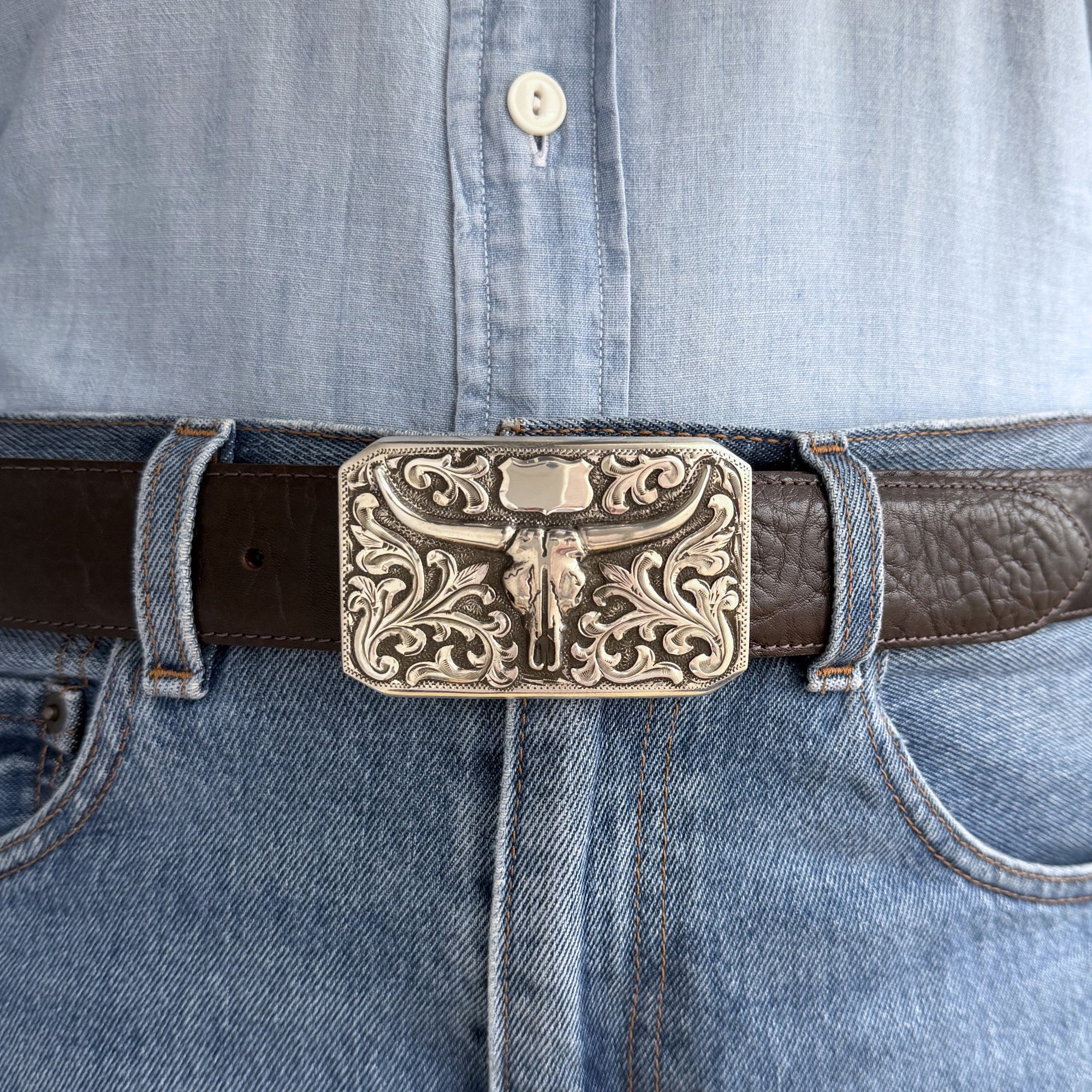 Rectangle Bohlin buckle with overlaid skull, shield and scrolls on brown belt. Model has blue shirt and blue denim shirt.