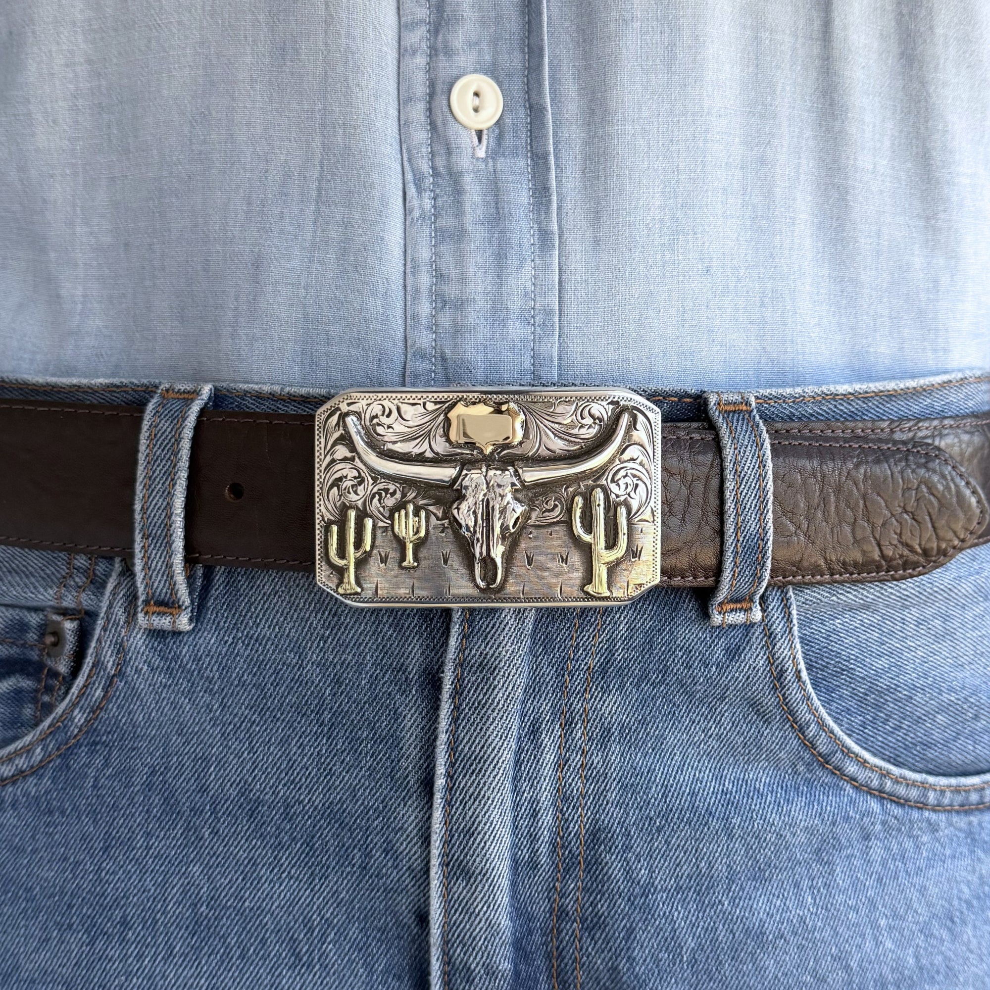 rectangular silver buckle with silver longhorn skull, green gold cacti and yellow gold shield at the top on brown belt.