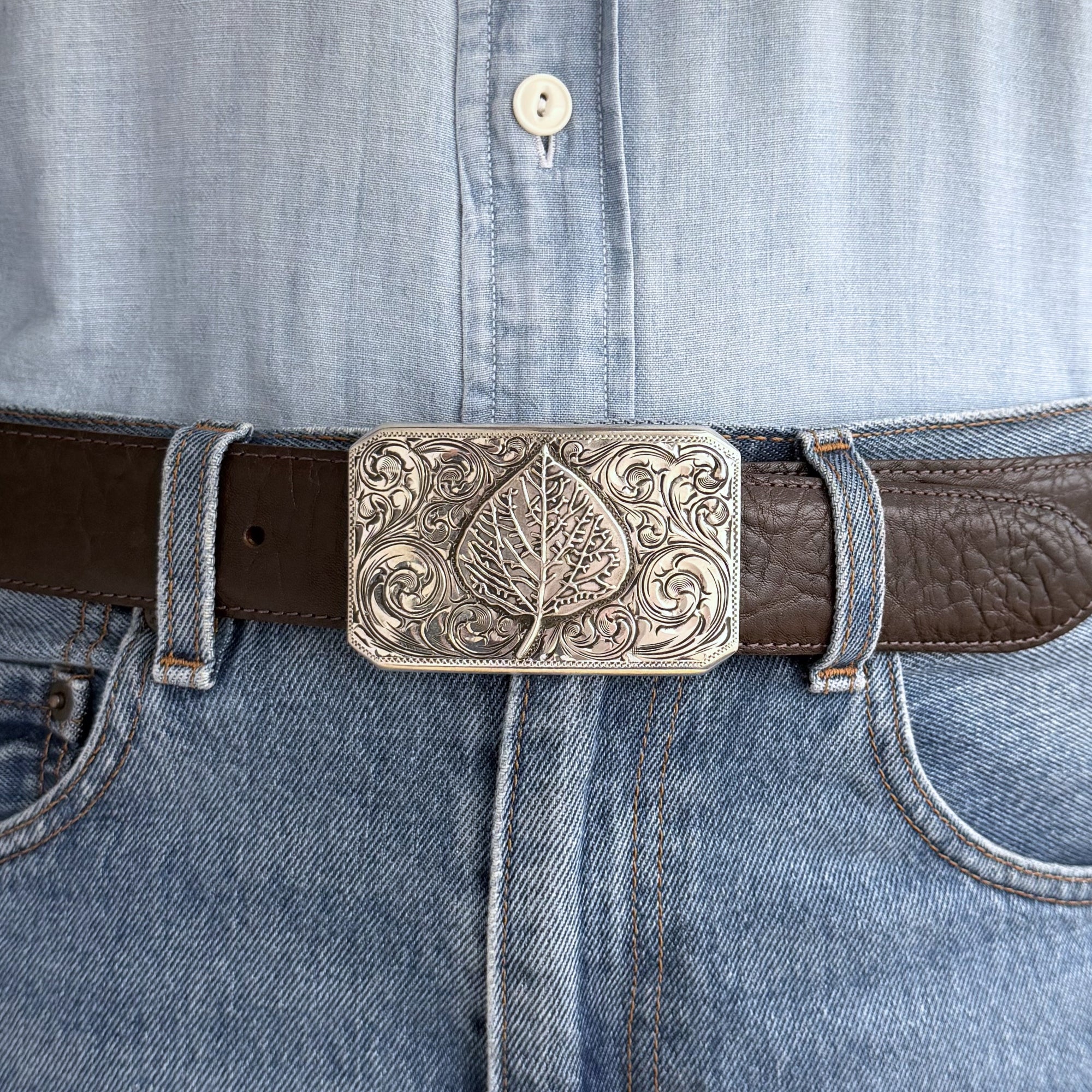 Rectangle Bohlin Belt buckle with engraved background and overlaid sterling silver aspen leaf on brown belt with jeans and a blue shirt.