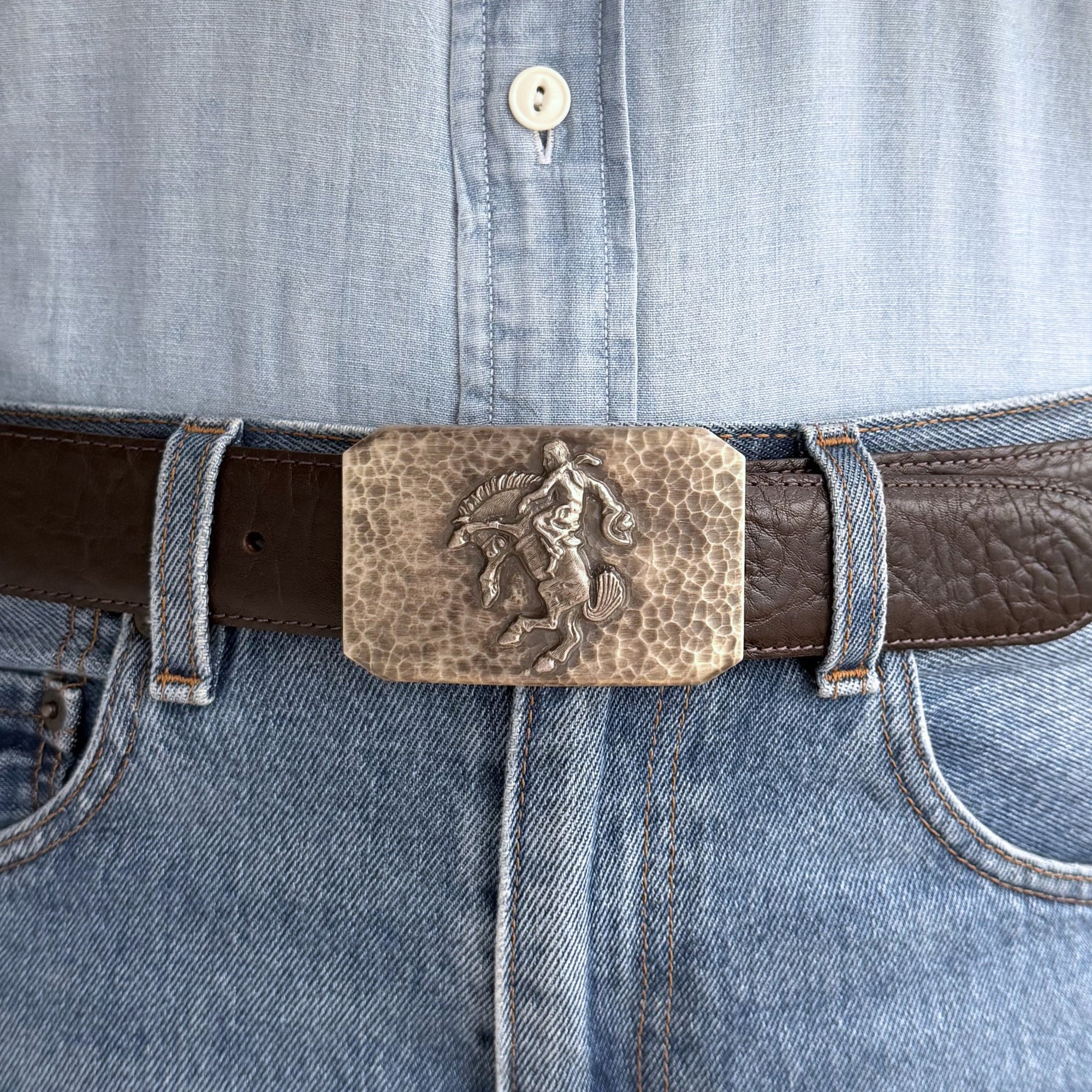 Hammered bucking horse buckle on brown belt, blue jeans and blue shirt