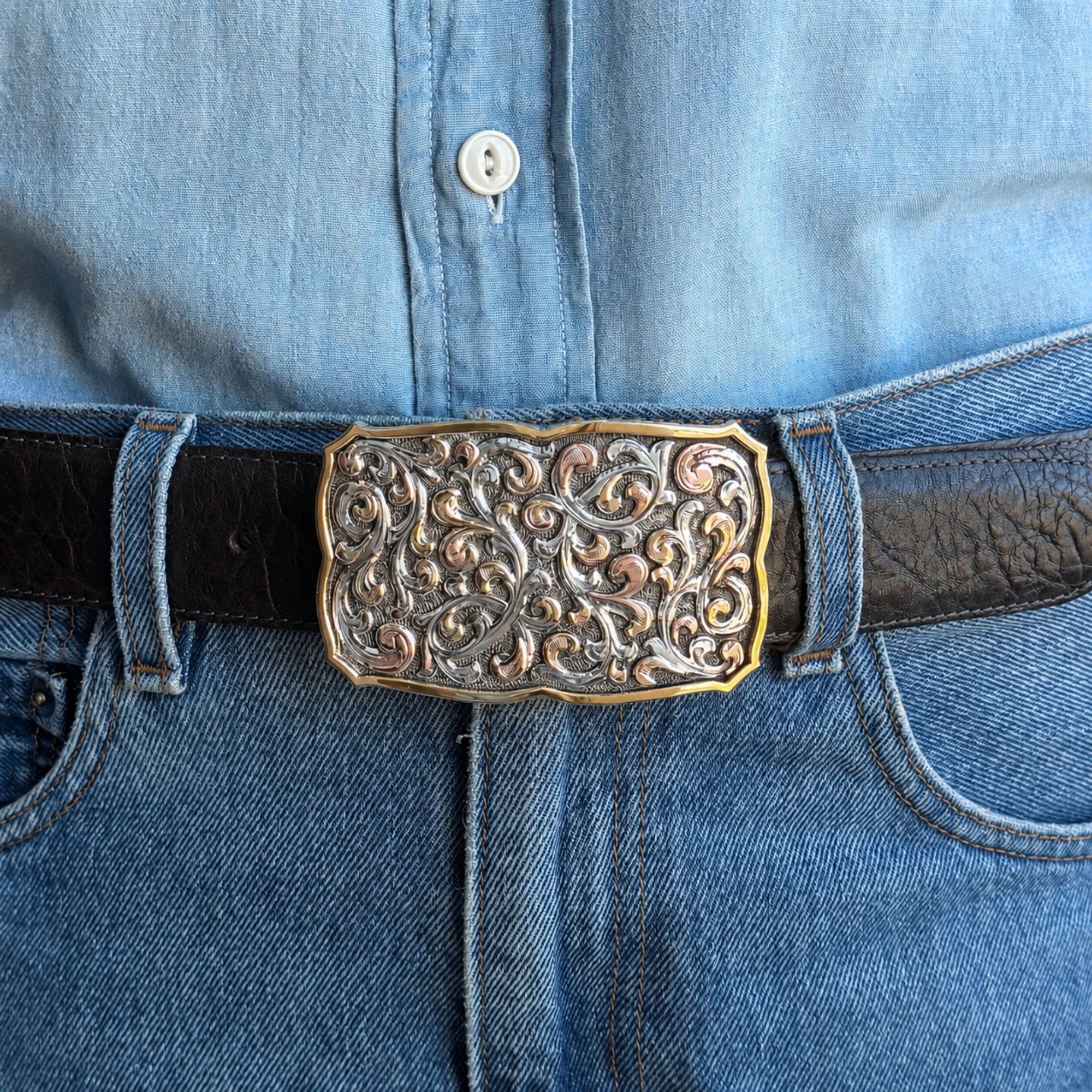 A close-up of a western-style belt with an ornate, engraved silver-toned buckle featuring gold-toned highlights, worn with a black leather belt over blue denim jeans and a light blue denim shirt.