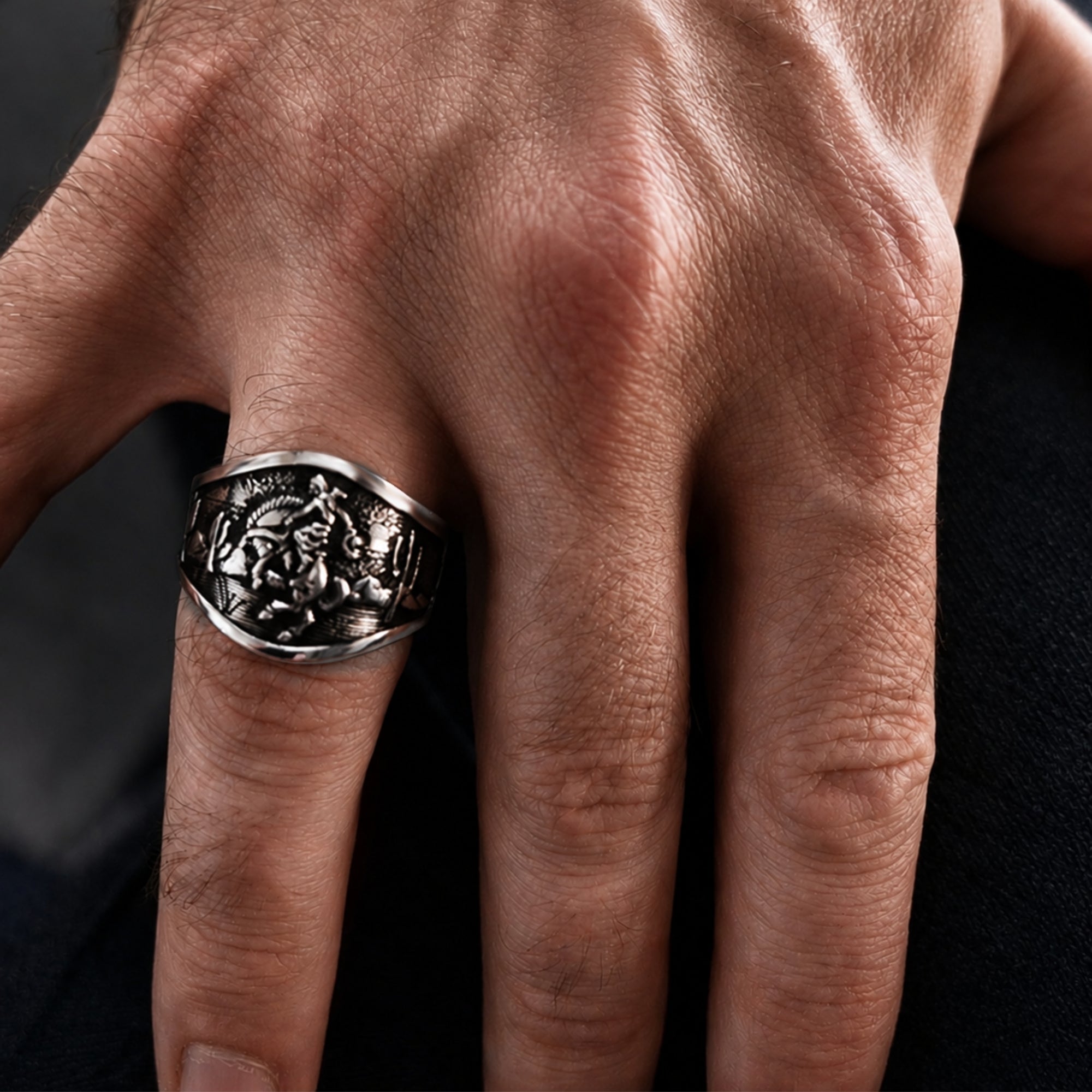 A close-up of a hand wearing a large silver-toned signet ring with an embossed floral or scrollwork design on the face.