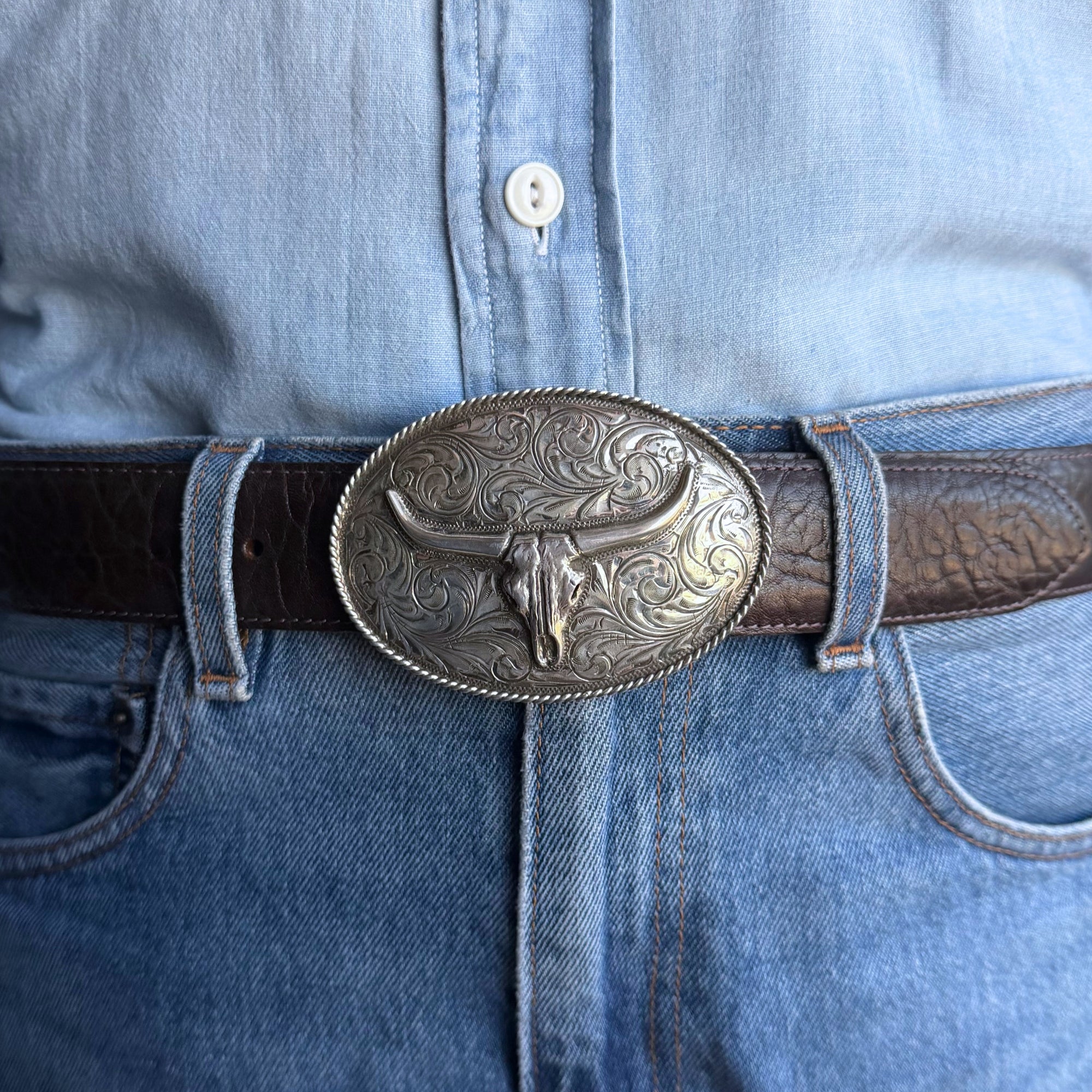 A close-up of a person wearing a dark brown leather belt with a large oval silver buckle featuring an overlaid skull with hand engraved scrolls and a twisted rope edge 