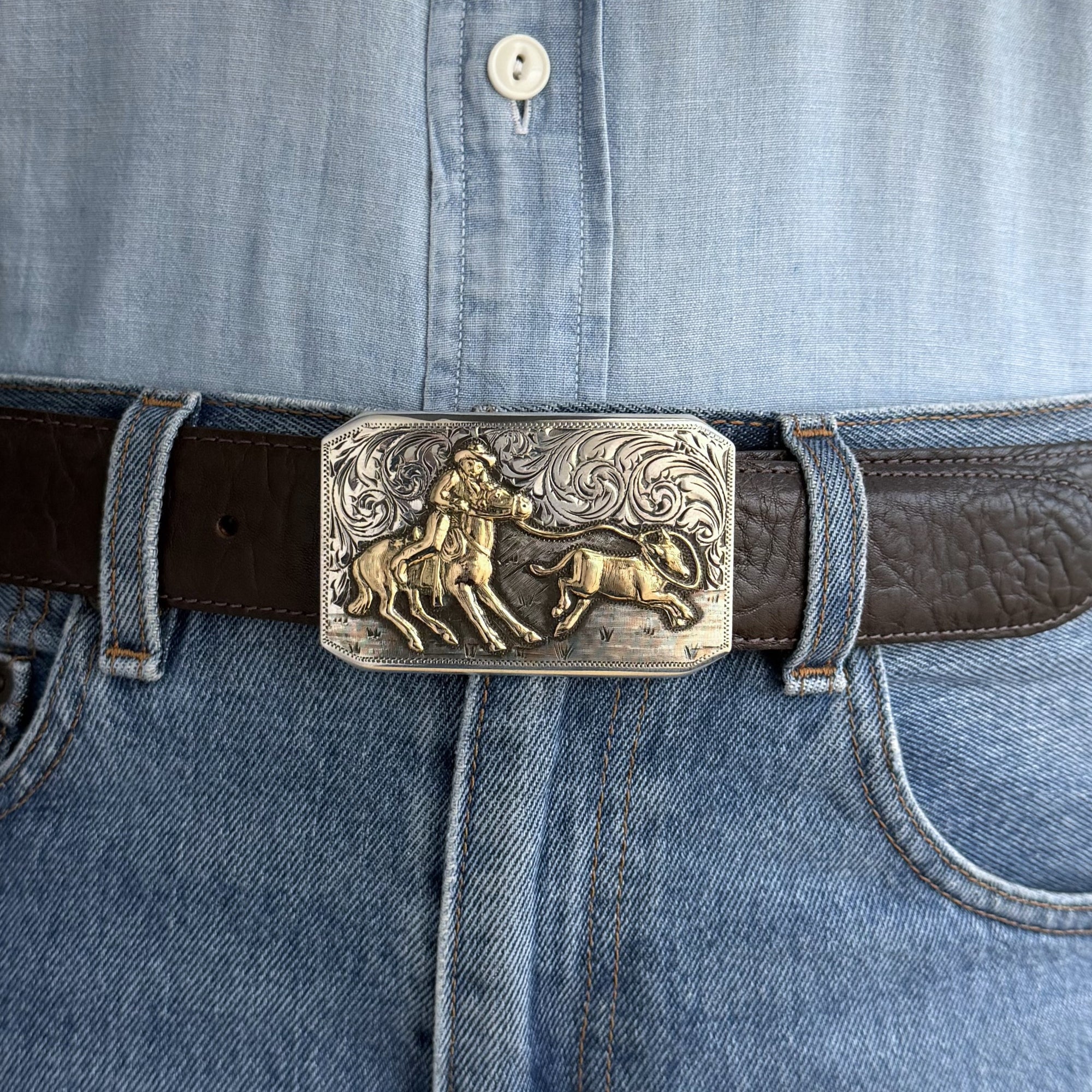 rectangle Bohlin buckle with engraved background and 14kt gold calf roper on brown belt. Blue shirt and Jeans also in the picture.