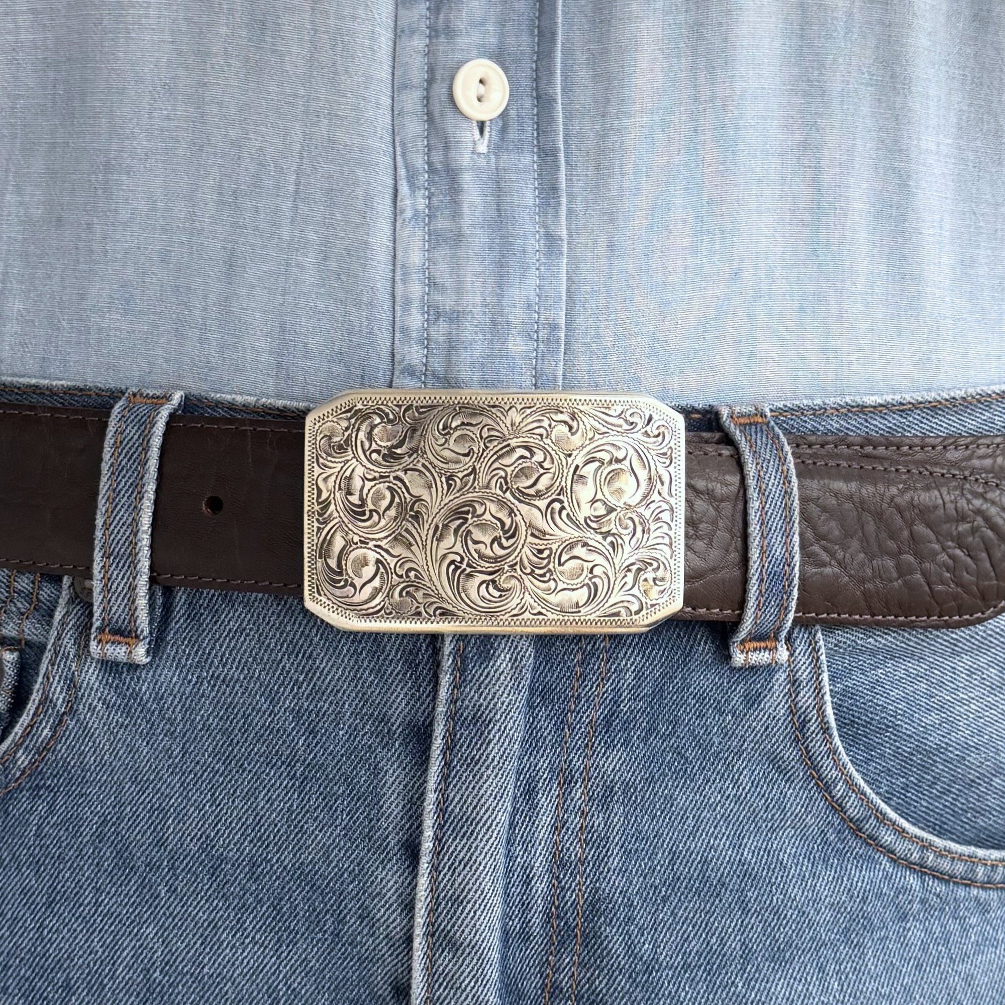 Fully engraved rectangular belt buckle on brown belt, jeans, and blue shirt 
