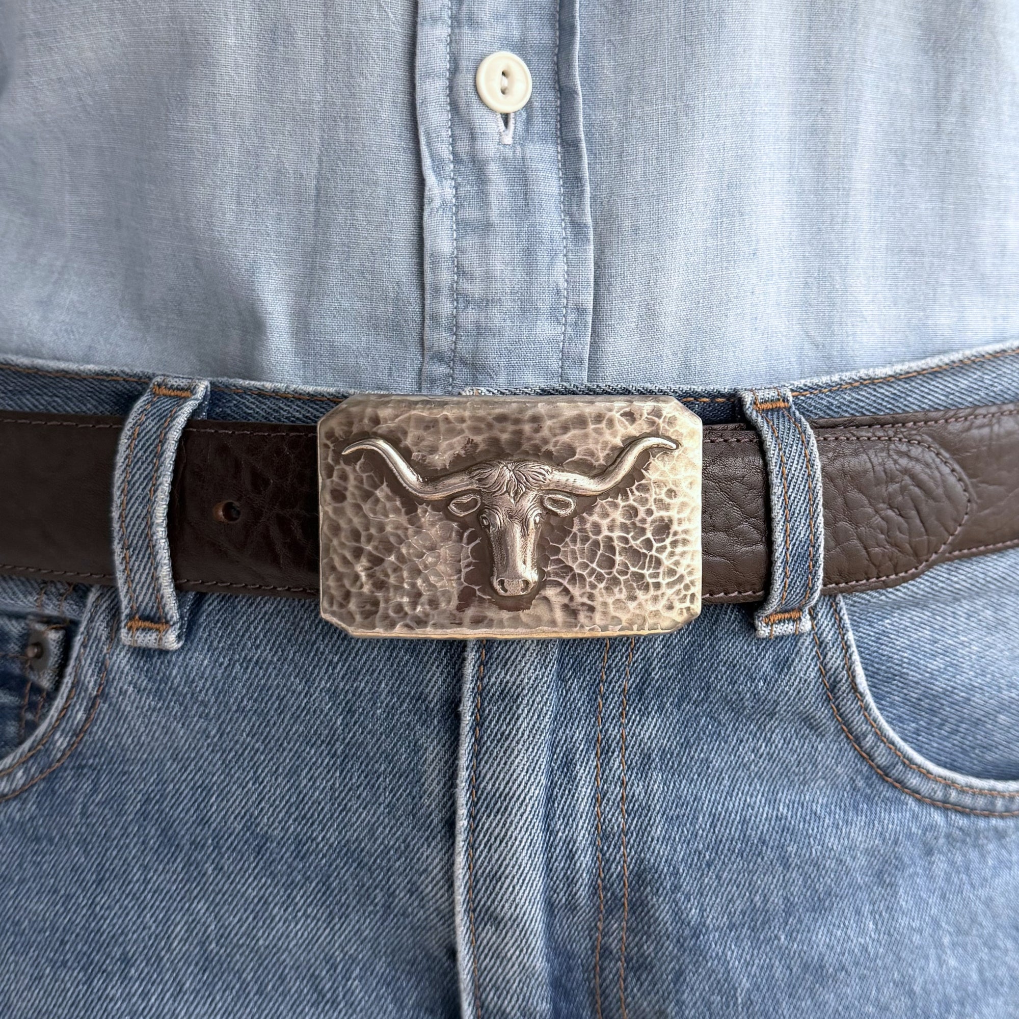 Brown belt with longhorn buckle worn on a light blue denim shirt.