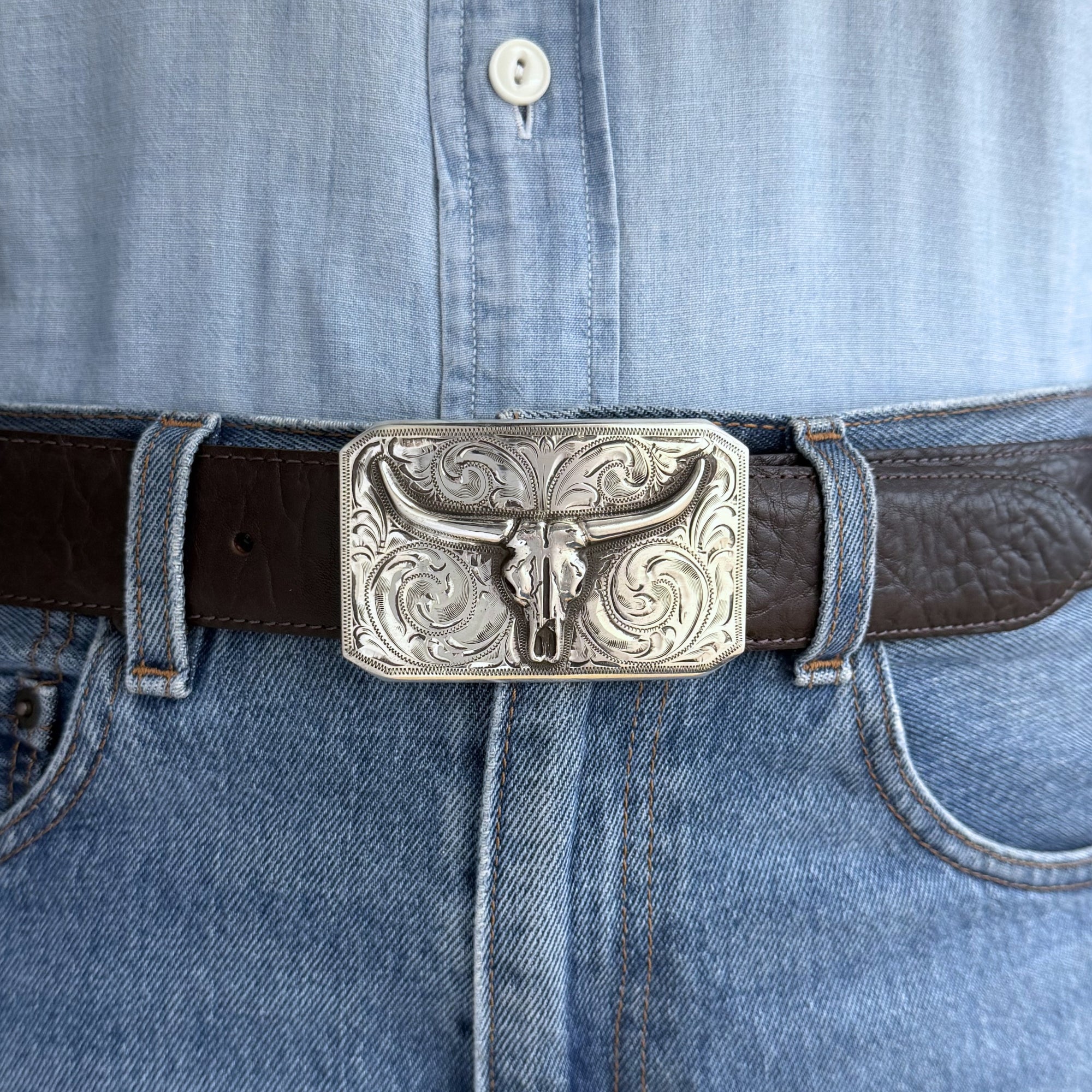 Close-up of a belt with a silver longhorn buckle on blue jeans.