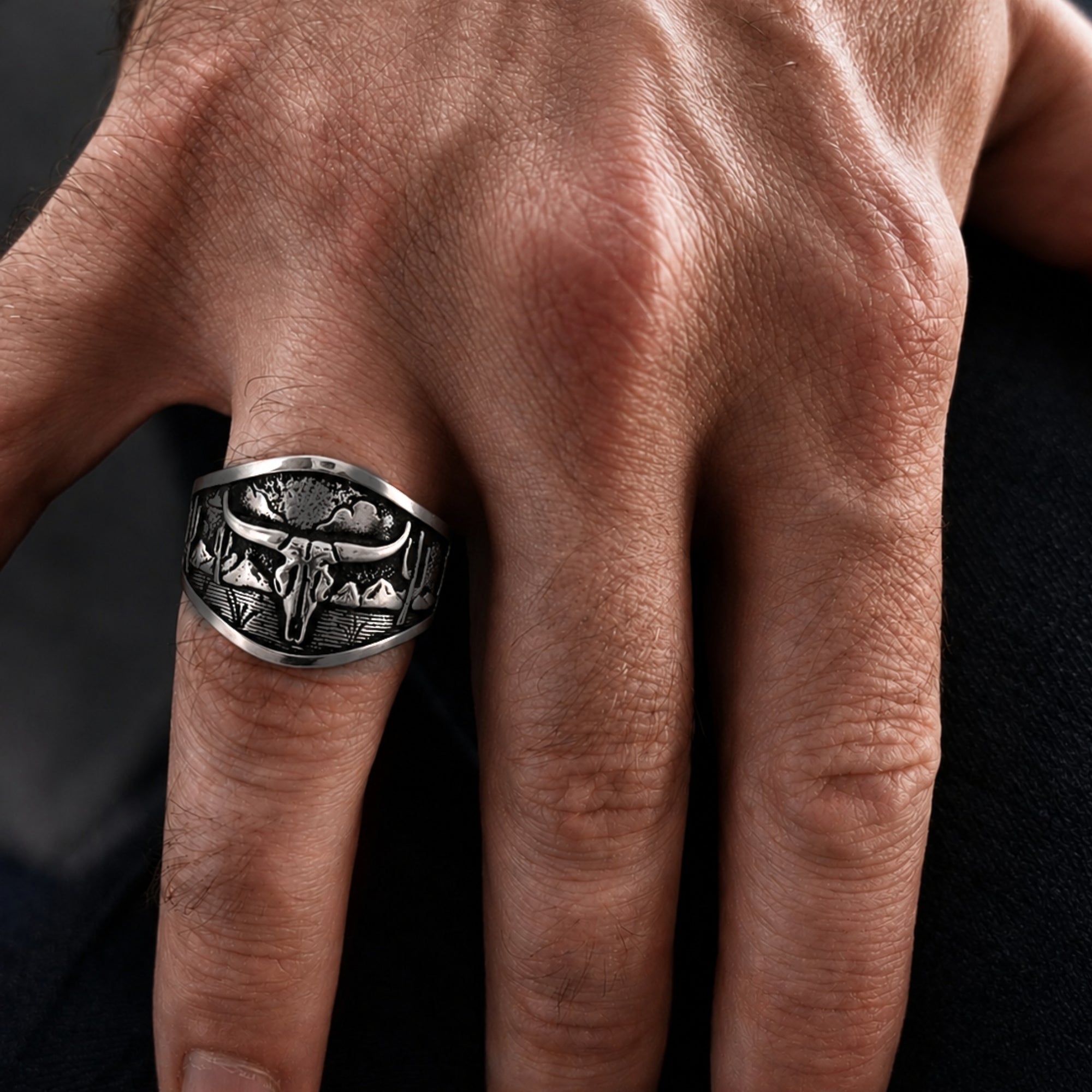 A close-up of a hand wearing a wide silver-toned ring with an engraved skull and mountain motif.