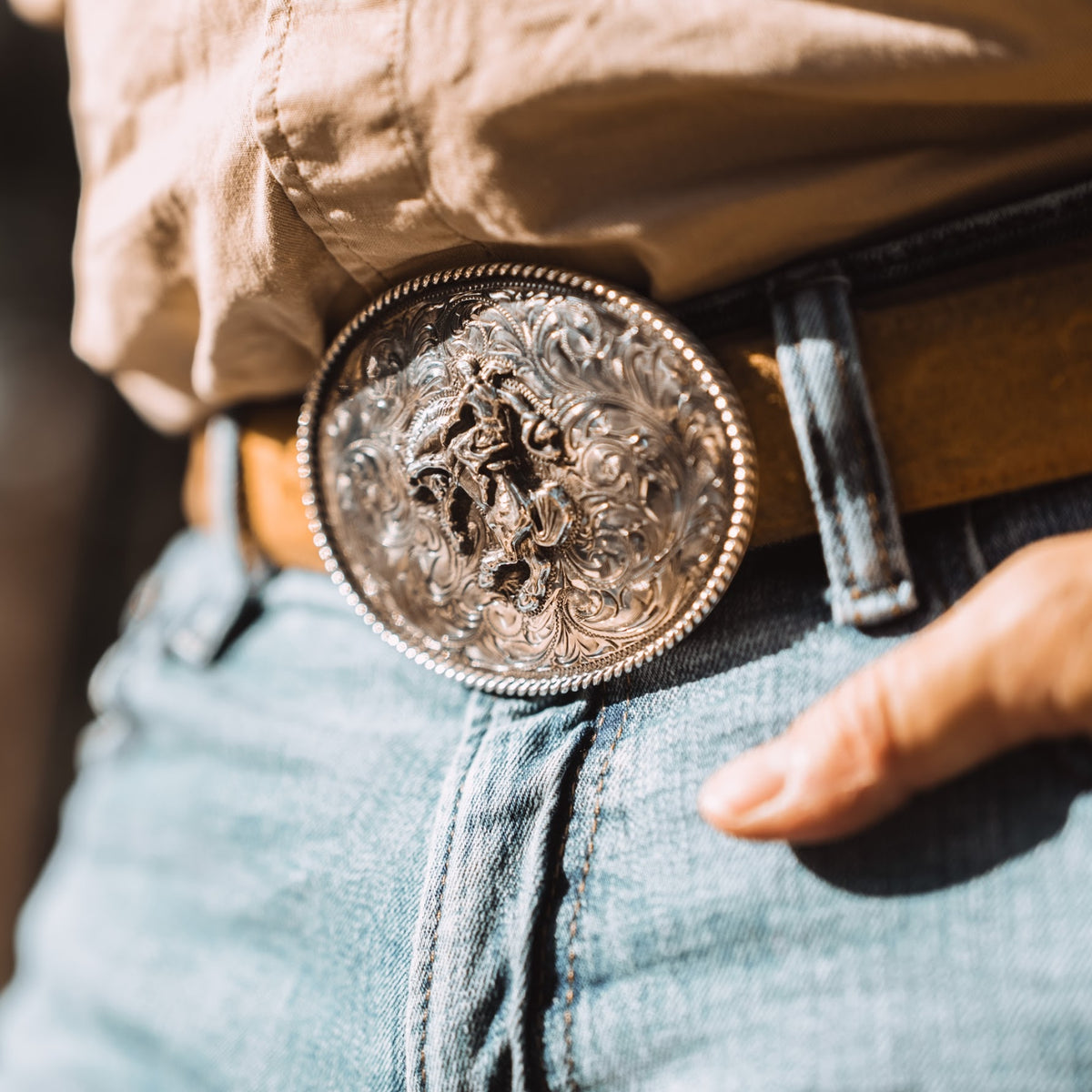 Oval hand fabricated belt buckle with bucking horse and hand twisted rope edge. Worn on brown belt with tan shirt and jeans.