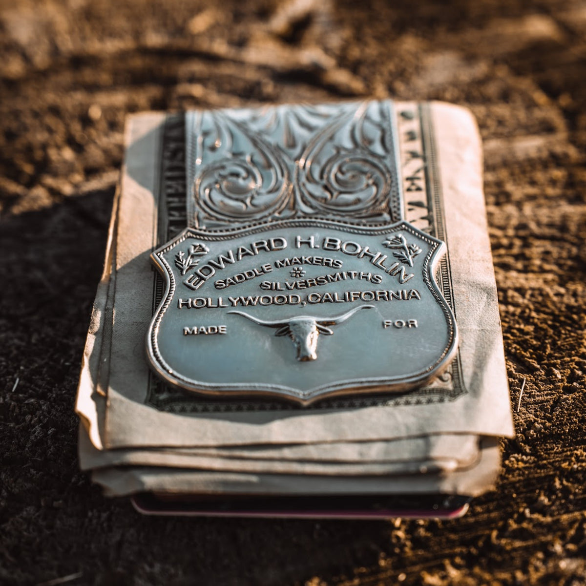 A vintage embossed silver money clip photographed outdoors on dark textured ground with warm sunlight and shadows.